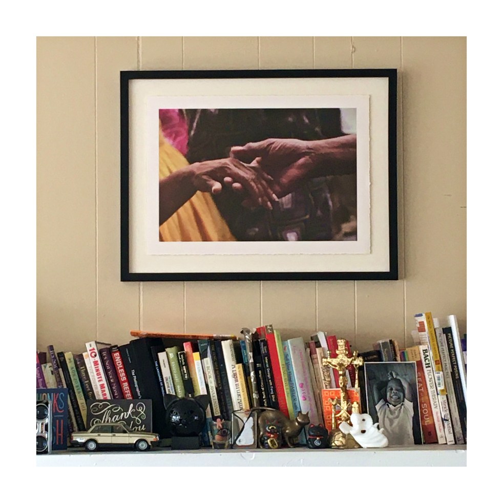 Image shows a shelf with many books and on top a framed picture of two hands.