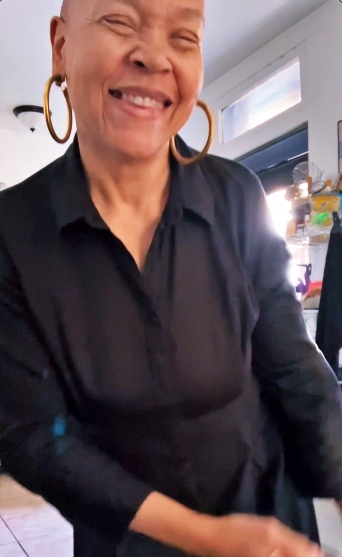 A smiling person with short hair, wearing a black shirt and large hoop earrings, standing in a well-lit indoor setting.